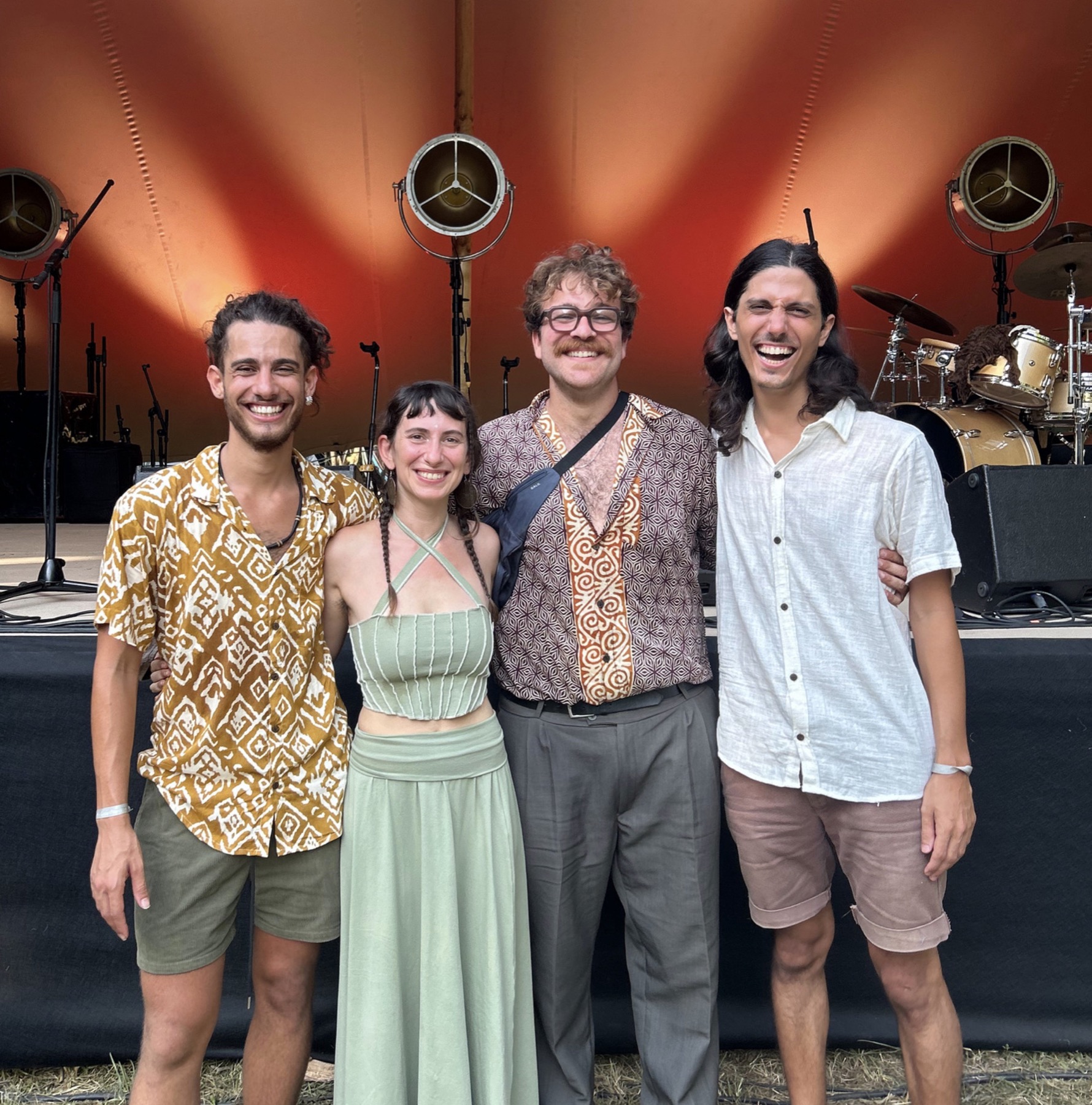 The four band members laughing together in front of a festival stage lit in orange